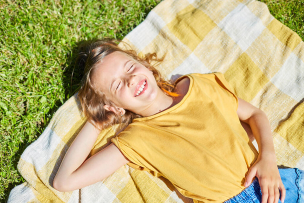 Child lying on the blanket, on the grass in the sun day, little girl take sunbathes on backyard of the house on a sunny summer day, summer time vacation. - Photo, Image