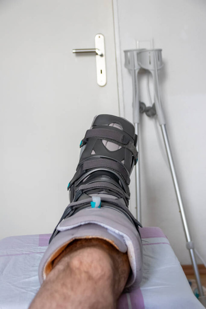 L'homme européen après l'opération de rupture du tendon d'Achille est de retour à la maison avec une chaussure de physiothérapie spéciale et des béquilles pour la récupération à la maison avec des médicaments sains analgésiques médicaments contre la jambe blessante - Photo, image