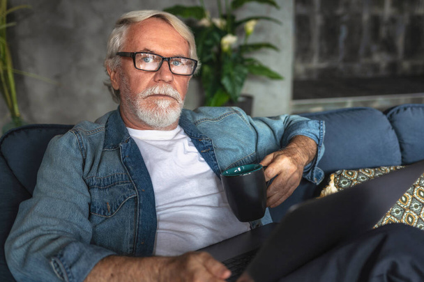 Portrait of senior business man with a cup of coffee and a laptop sitting on the sofa at home or in a modern office looking directly at the camera - 写真・画像