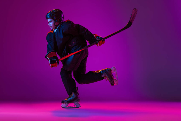 Portrait of child. hockey player training isolated over purple background in neon light. Childhood hobby - 写真・画像