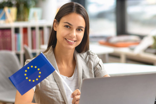 Beautiful Caucasian woman student with EU flag sitting at table with laptop and smiling while looking at camera. EU education concept - Foto, Imagem