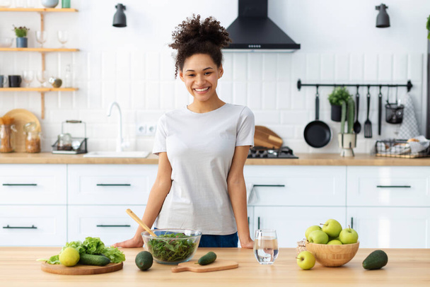 Healthy diet eating. African American young female preparing salad in home kitchen. Beautiful woman cooking healthy food, breakfast or dinner. Looking at the camera and smiles friendly - Foto, afbeelding