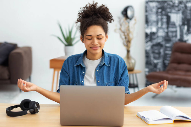 Young African American woman sitting at the office desk with a laptop meditating eyes closed thinking of good things and focusing on positive feelings and emotions - 写真・画像