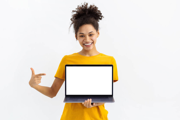 Portrait of young African American woman with laptop on white background. Beautiful female points her finger at a blank laptop screen, looking at the camera and smiling - Фото, зображення