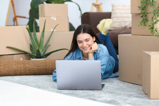 Moving to a new house, rental housing. Happy young caucasian woman using laptop computer to search and order a transportation service and movers to move to a new home - Photo, Image