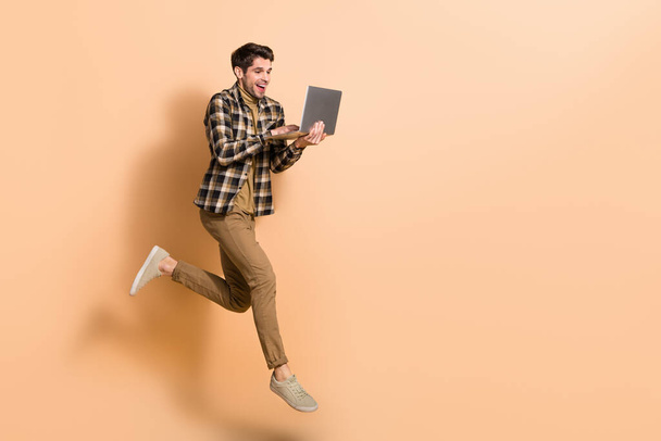 Full length body size view of attractive cheery skilled guy jumping using laptop copy space isolated on beige pastel color background - Photo, Image