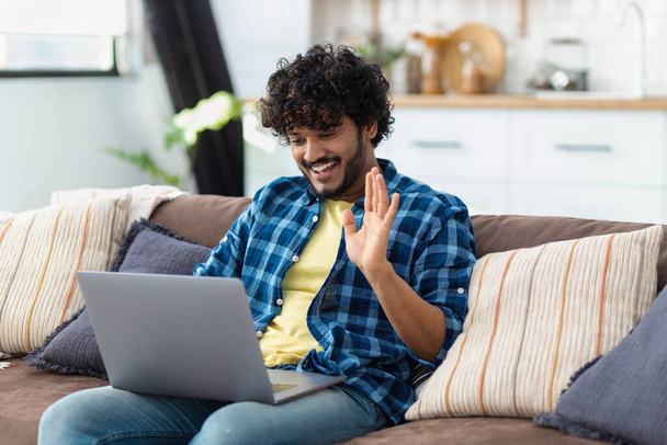 Smiling young Indian man relaxing on the couch using video call to chat with friends online at home, waving hand and smile friendly - Φωτογραφία, εικόνα