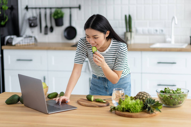 Cooking healthy food concept. Young asian woman watching recipe online using laptop computer while standing at home kitchen - Photo, Image