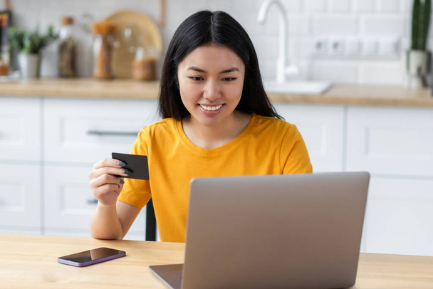 Happy smiling young Asian woman with laptop computer holding credit card sitting at home, shopping online, ordering food, booking tickets on website. Looking at the camera and smiles friendly  - Foto, Imagem