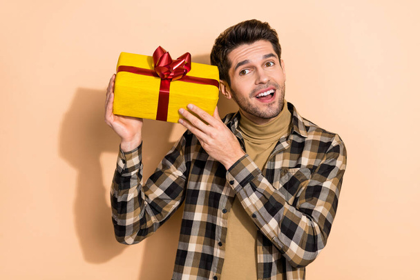 Portrait von attraktiven fröhlichen verträumten Kerl hält Geschenkbox erraten, was sich im Inneren isoliert über beige Pastellfarbe Hintergrund - Foto, Bild