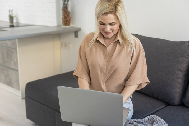 Nice beautiful blonde lady works at the notebook sit down on the sofa at home - check on oline shops for cyber monday sales - technology woman concept for alternative office freelance - Foto, Imagem