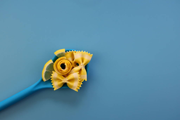 Żółty makaron farfalle, plastry i skórka cytryny w niebieskiej łyżeczce na niebieskim tle. Pasta cytrynowa Farfalle - Zdjęcie, obraz