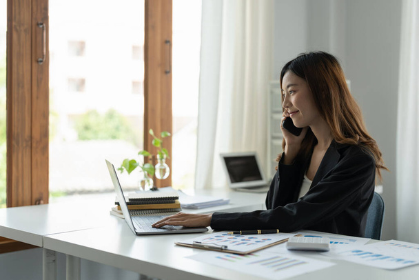 Aziatisch zakenvrouw praten op mobiele telefoon werken op laptop voor financieel in modern kantoor. - Foto, afbeelding
