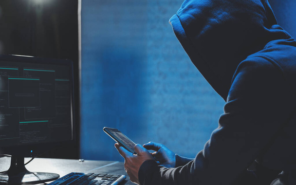 Накладний хакер у капюшоні, що працює на комп'ютері та мобільному телефоні, друкує текст у темній кімнаті, анонімний хакер використовує шкідливі програми з мобільним телефоном, щоб зламати пароль, особисті дані крадуть гроші з банку. кібернетичний
 - Фото, зображення