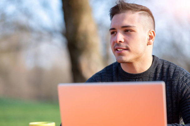 Portrait of attractive young office worker in casual wear, smiling in a break moment. He works remotely with the laptop from a table in the park, remote working concept - Photo, Image