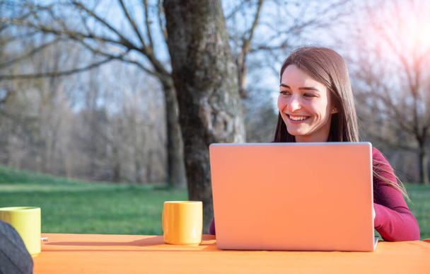 Retrato de mujer joven trabaja remotamente en un parque en un día soleado agradable. Ella usa un portátil mientras habla entre sí. En la mesa hay tazas. Concepto de trabajo remoto al aire libre. - Foto, Imagen