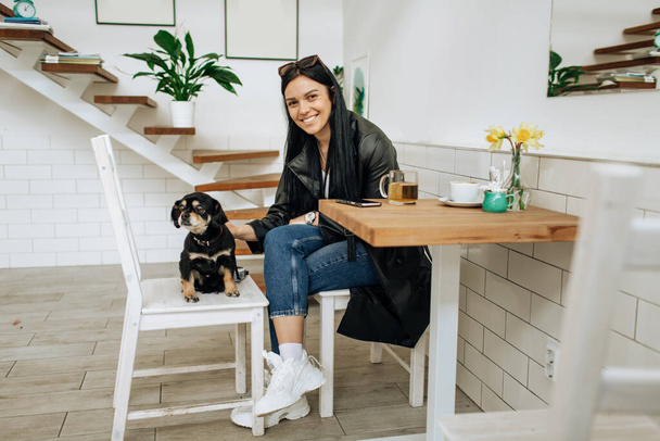 An optimistic woman in a gray suit smiles and plays with a dog in a cafe. Pretty woman in stylish coat posing. High quality photo - Foto, Imagen