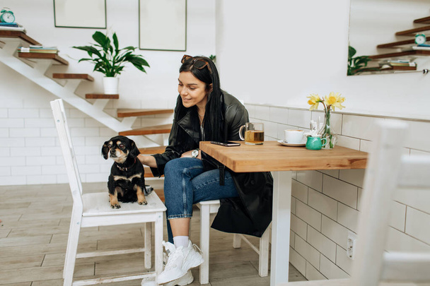 An optimistic woman in a gray suit smiles and plays with a dog in a cafe. Pretty woman in stylish coat posing. High quality photo - Фото, изображение