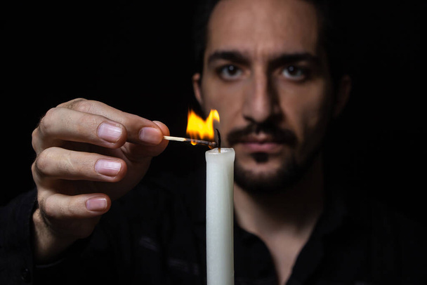 Ένας άντρας ανάβει ένα κερί σε μαύρο φόντο. Ανάψτε ένα κερί ως σημάδι μνήμης και θλίψης. μικρή φωτιά κεριών - Φωτογραφία, εικόνα