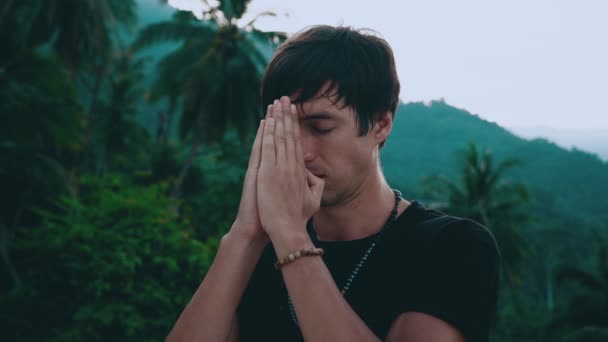 Religious young man holding hands in namaste gesture standing on beautiful jungle nature with eyes closed. Male is praying brings his hands to his face and begins to say prayer. - Footage, Video