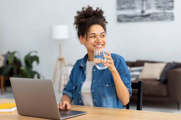 Retrato de mujer freelancer o empleada con vaso de agua dulce sentada en el lugar de trabajo, co-working. Mujer afroamericana sonriente usando computadora portátil, trabajando en casa. aprendizaje, educación en línea - Foto, imagen