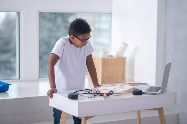 A boy in a white tshirt standing at the table with cables and laptop - 写真・画像