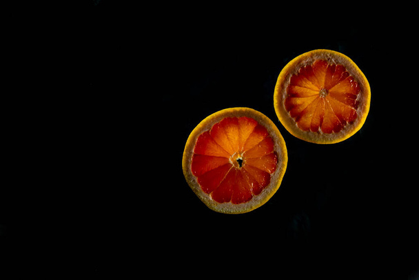 Κομμένο γκρέιπφρουτ μισοαπομονωμένο σε μαύρο φόντο. Ροζ, πορτοκαλί Φρέσκα γκρέιπφρουτ εσπεριδοειδών, φέτα με ξύσμα, δραματική ελαφριά υφή, πολύχρωμα κομμένα φρούτα ως φόντο φωτισμένο, αντίγραφο χώρου - Φωτογραφία, εικόνα
