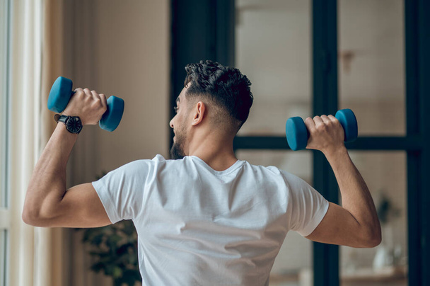 Young dark-haired man exercising with dumbbells and looking contented - Foto, immagini