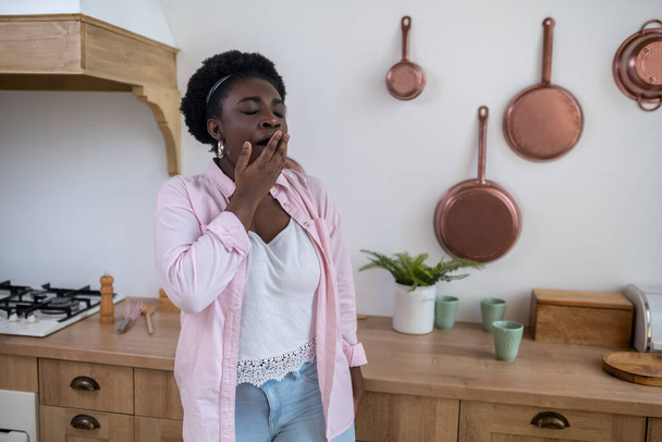 A dark-skinned woman standing in the kitchen and yawning - Фото, изображение
