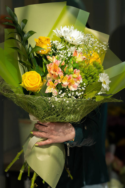 Close-up de um belo buquê de flores diferentes em um pacote verde claro brilhante na mão de uma mulher. Presente, salão de flores. Foto vertical - Foto, Imagem