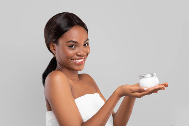 Blij mooie jonge Afrikaan amerikaanse dame met glanzend haar in handdoek houden pot van crème, geïsoleerd op grijze achtergrond - Foto, afbeelding