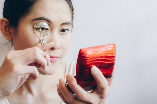 Portret van een jonge Aziatische vrouw die naar de spiegel kijkt met een wimperkruller. Eyelash curler is een met de hand bediend mechanisch apparaat voor het krullen van wimpers voor cosmetische doeleinden.  - Foto, afbeelding