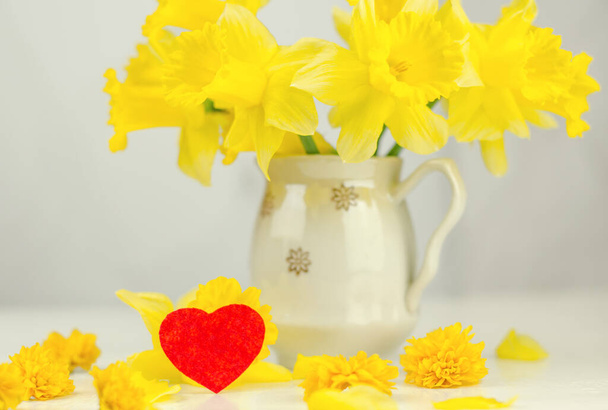 Spring postcard. Vase with a bouquet of daffodils and a red heart on a light background, selective focus - Photo, Image