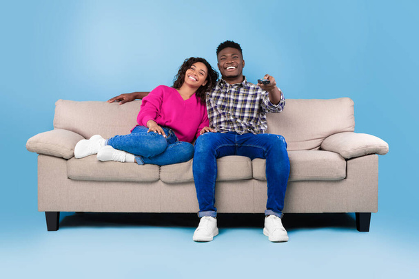 Excited young black couple with remote control sitting on couch and watching TV against blue studio background - Photo, image