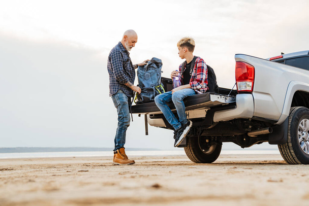 White mid aged father and his preschooler son sitting on a back of a car parked at the beach holding camping gear - Foto, immagini