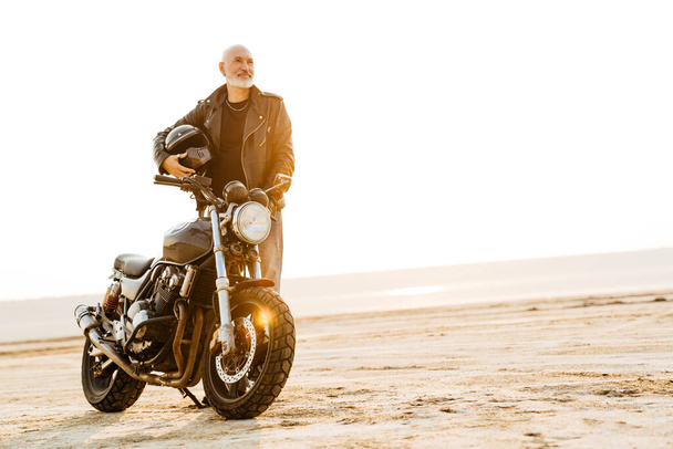 Bold senior man in leather jacket posing with motorcycle outdoors on summer day - Valokuva, kuva
