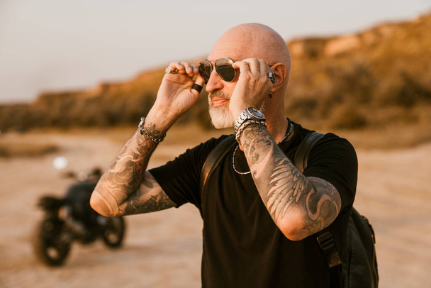 Bold senior man wearing eyeglasses posing with motorcycle outdoors on summer day - Foto, Bild