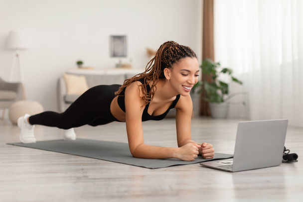 Μαύρη γυναίκα κοντά lap-top στέκεται σε σανίδα άσκηση στο σπίτι - Φωτογραφία, εικόνα