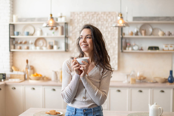 Start in den Tag. Porträt einer fröhlichen Frau, die den morgendlichen Kaffee genießt, die Tasse in der Hand hält und in der Küche steht - Foto, Bild