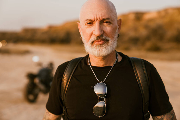 Bold senior man wearing t-shirt posing with motorcycle outdoors on summer day - 写真・画像