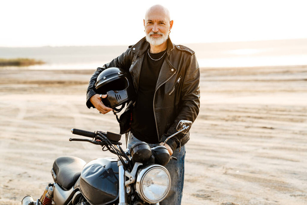 Mature bold man holding helmet while posing with motorbike outdoors - Foto, Bild