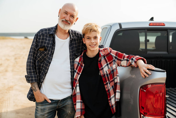 Lächelnder Vater und sein Sohn stehen lachend am Kofferraum eines Autos am Strand - Foto, Bild
