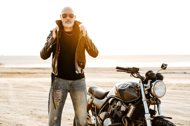 Bold senior man in leather jacket posing with motorcycle outdoors on summer day - Foto, Imagem