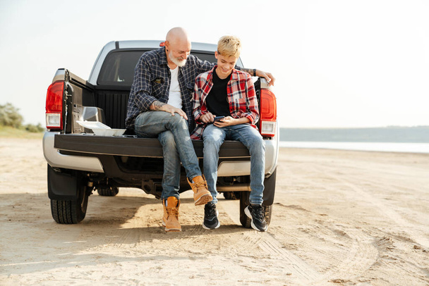 Smiling father and his son sitting in a car trunk on a back at the beach having lunch talking - Foto, Bild