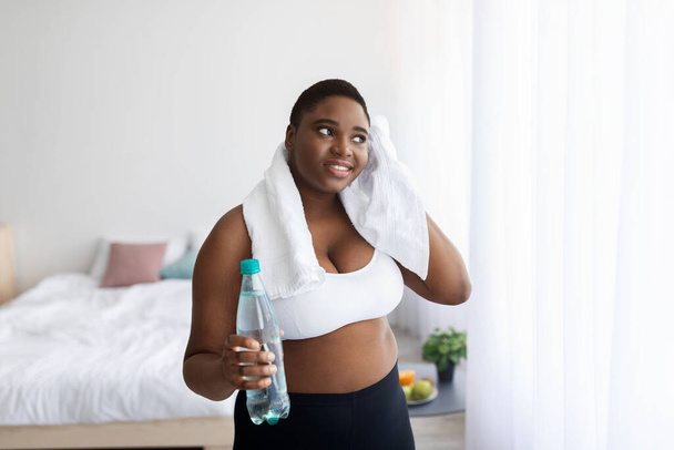 Grande taille jeune femme afro-américaine avec bouteille d'eau essuyant la sueur avec une serviette après l'entraînement à la maison - Photo, image