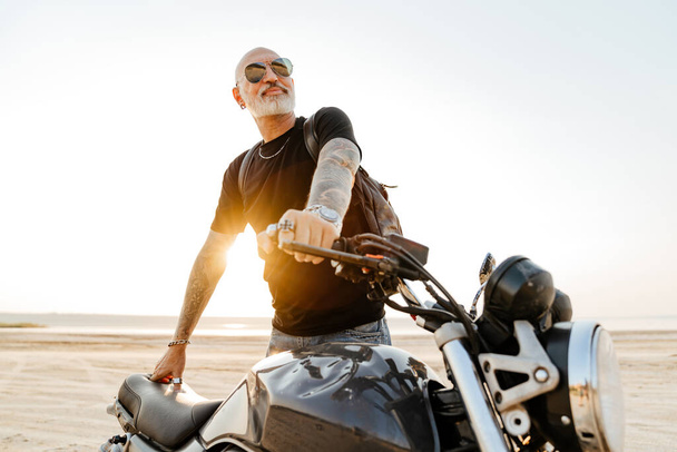 Bold senior man in sunglasses posing with motorcycle outdoors on summer day - 写真・画像