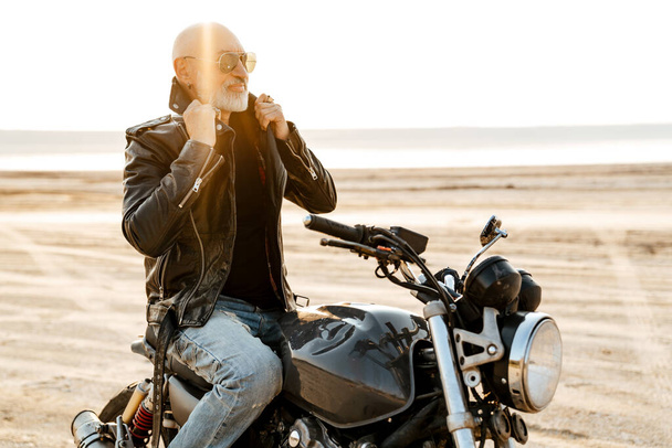 Bold senior man wearing leather jacket posing on motorcycle outdoors on summer day - 写真・画像