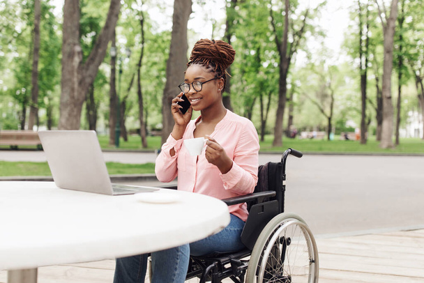 Jeune femme noire en fauteuil roulant travaillant en ligne, en utilisant un ordinateur portable, parlant sur smartphone et dégustant du café au café extérieur - Photo, image