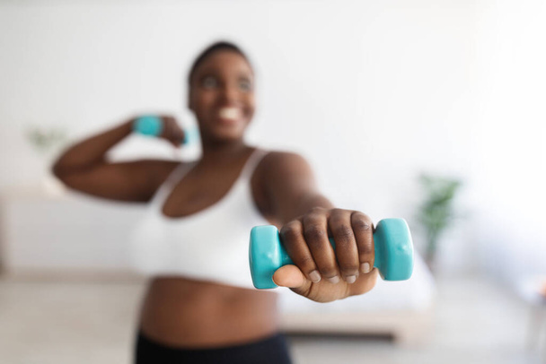 Συν μέγεθος νεαρή μαύρη γυναίκα κάνει ασκήσεις με αλτήρες, ενίσχυση του σώματός της στο σπίτι, επιλεκτική εστίαση - Φωτογραφία, εικόνα