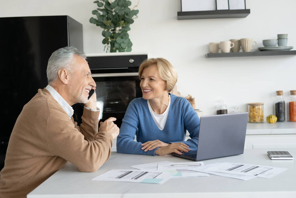 Happy elderly married couple laughing, talking on smartphone with family, sitting on kitchen, checking utility bill. Husband and wife call to bank support, regarding the last mortgage payment - Photo, Image